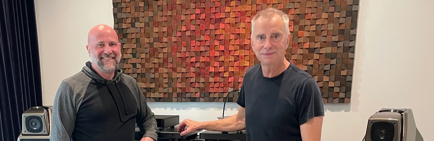 Two people beside a hi-fi system in a Sound Sanctuary Austin listening room, with floorstanding loudspeakers either side and a wooden mosaic artwork behind them.